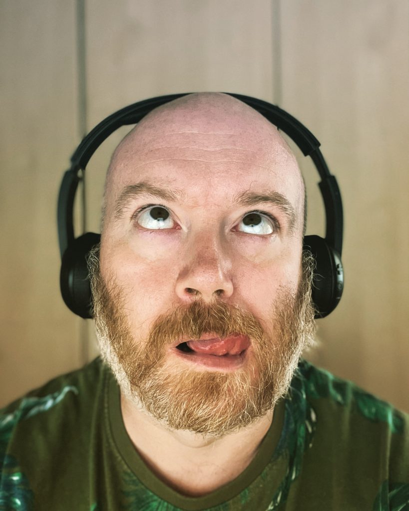 A close-up selfie of a cis, white bald man with a ginger beard. He's wearing black headphones and a green flowery T-shirt. His eyes are rolled to the ceiling and his tongue is sticking out.