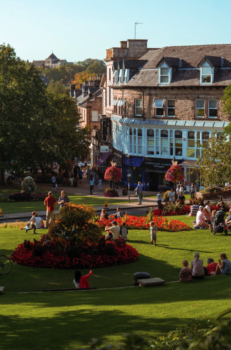 A park with lush green grass, multiple flower beds on a sunny day. People are sitting, relaxing and enjoying the day. In the background are Victorian buildings, possibly some shops.