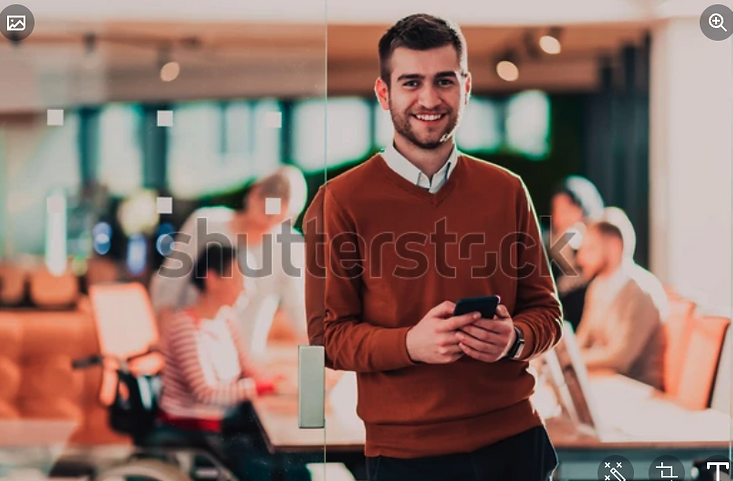 A man standing and leaning against a glass door in an office holding a phone. It is the same man from the earlier image, but not in a wheelchair.