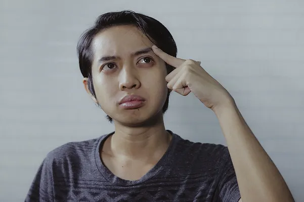 A man of Asian origin looks upwards with a finger on his forehead looking confused.