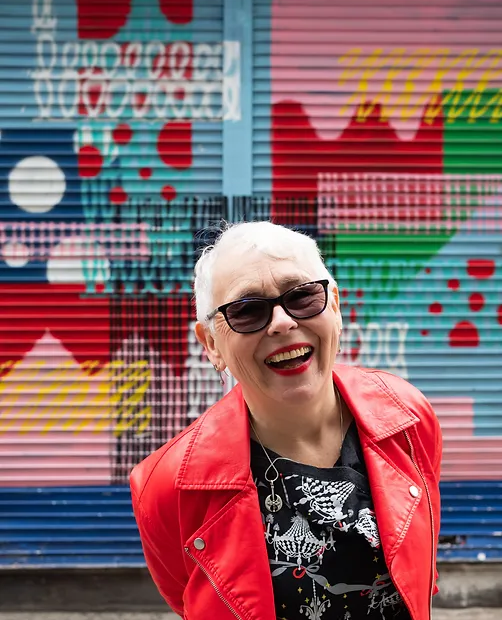 A woman with short, white hair is standing in front of a multicoloured and vibrant shop shutter. She is wearing a bright red, leather coat with a black top and sunglasses. She has a wide, happy smile.