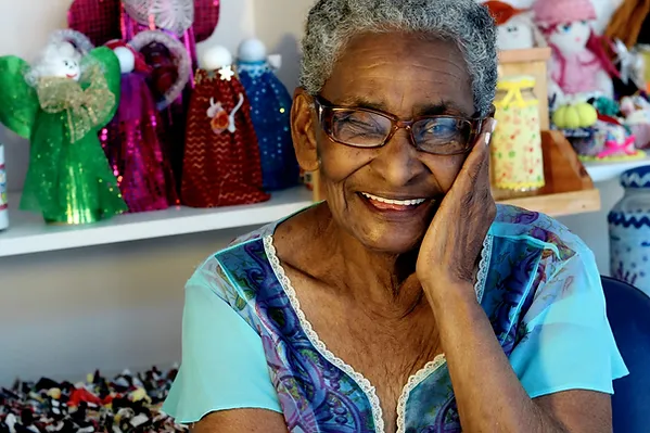 A black woman with grey hair and glasses wears a blue top. She is smiling at the camera and is resting her cheek on her hand. Behind her are multicoloured dolls on shelves.