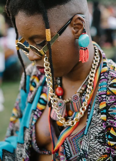 A black woman in close up with multiple pieces of jewellery, necklaces and earrings on. She is wearing sunglasses which are attached to large gold chains that go around her neck. She is wearing a multicoloured shirt and has two dreadlocks and a shaved head.