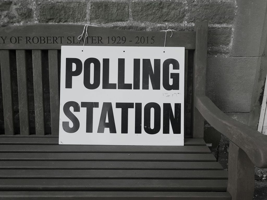 a black and white photo of a sign saying Polling Station on a bench.