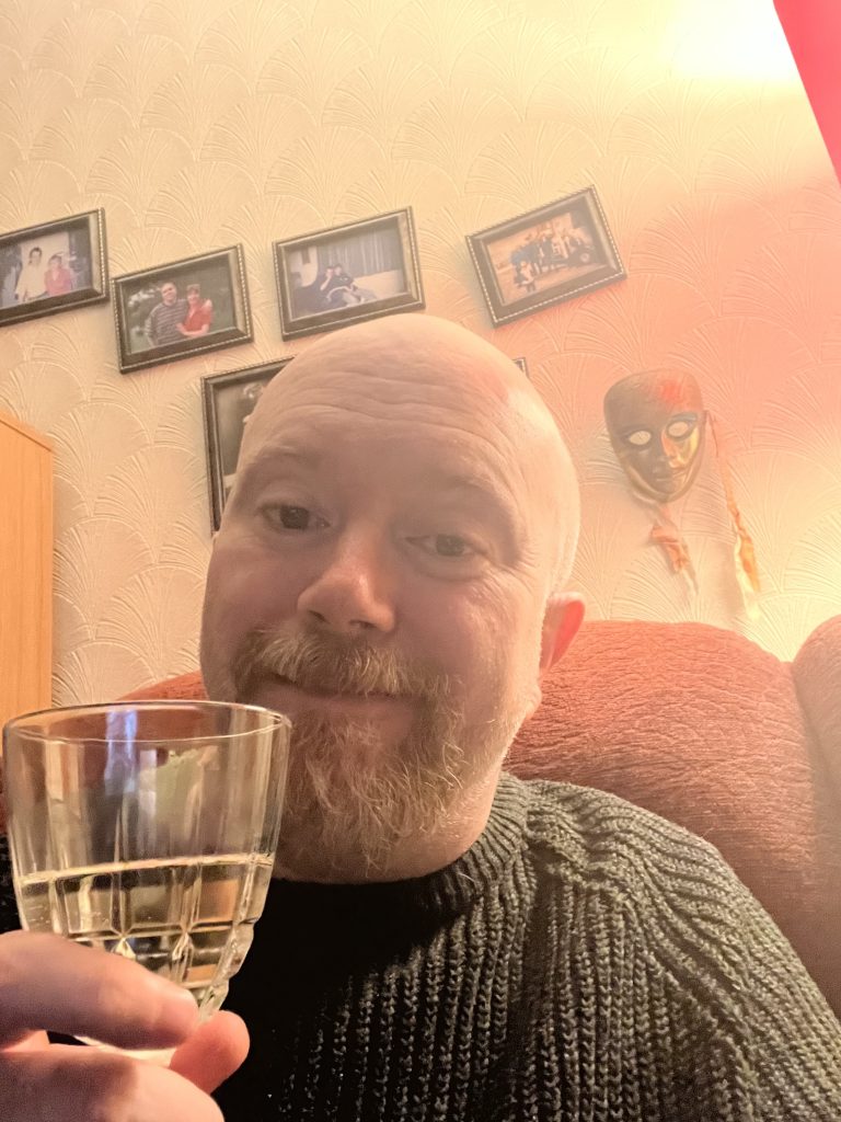 Peter, a cis white bald man with a beard holds a half empty wine glass while sitting on a red sofa with personal photos on the wall behind him.