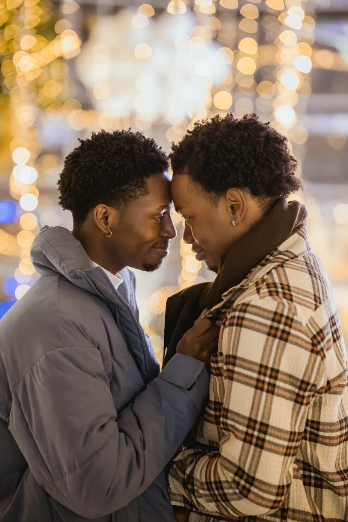 Two Black men gaze into each other's eyes, touching foreheads in an embrace of love. They both have an earring, and wear large winter coats.