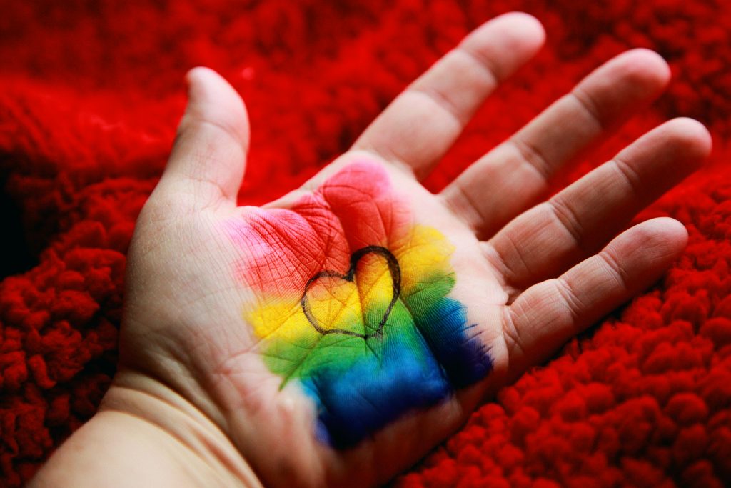 A white person's hand, palm facing up, with rainbow colours painted on the palm and a black outlined heart in the middle.